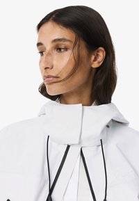 Giacca bianca impermeabile con colletto oversize, cordini regolabili e tasca superiore testurizzata con logo.