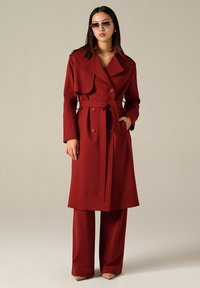 Trench-coat rouge à double boutonnage avec une ceinture à nouer, grands revers et poches latérales, associé à un pantalon large et des lunettes de soleil discrètes.