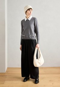 Grauer Pullover mit einem weißen Kragen, zwei vorderen Taschen mit Reißverschluss, weiten schwarzen Hosen und einer cremefarbenen Schultertasche. Schwarze Schuhe vervollständigen das Outfit.