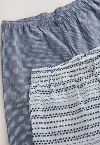Deux paires de pantalons avec taille élastique posées à plat, l'une à motif à carreaux bleu et l'autre blanche avec des lignes pointillées bleu marine.