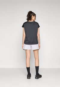 Camisa negra de manga corta, tela ligera, con dobladillo redondeado, combinada con pantalones cortos grises claros, calcetines negros y zapatos deportivos. Vista trasera.