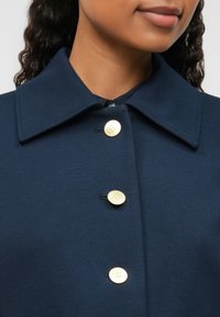 Zbliżenie na osobę noszącą granatowy płaszcz z szerokim kołnierzem i trzema widocznymi złotymi guzikami z przodu.