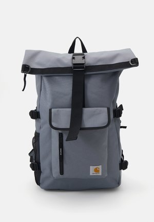 Mochila gris con tapa enrollable, bolsillo delantero con cremallera, correa negra con cierre de hebilla, bolsillo lateral de malla y parche con logo de Carhartt en la parte frontal.