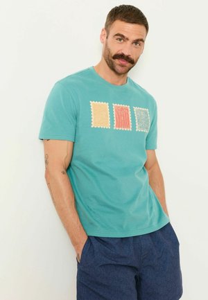 Mann mit Schnurrbart, der ein türkisfarbenes T-Shirt mit drei bunten Stempeldesigns und gemusterte marineblaue Shorts trägt, steht mit den Händen in den Taschen.