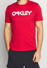 Czerwona bawełniana koszulka z krótkimi rękawami, z dużym białym logo "OAKLEY" na klatce piersiowej. Prosty, okrągły dekolt.