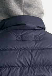 Gros plan du dos d'une doudoune bleu marine avec des coutures matelassées et une étiquette rectangulaire en tissu portant le nom de la marque GANT.