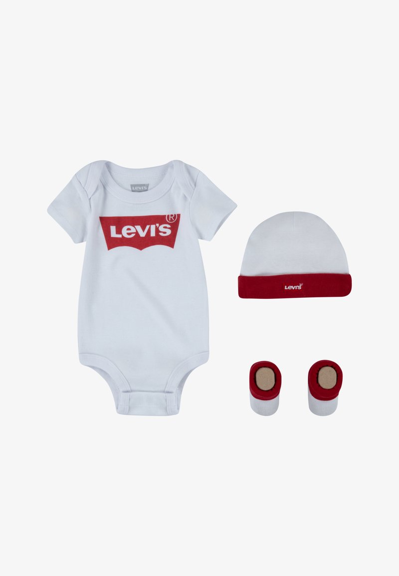 Balts kokvilnas kombinezons ar sarkanu Levi's logo, atbilstoša sarkaniem maliņu aksesuāriem, baltā cepurītē un zābaciņos, visi ar maigu, gludu tekstūru.