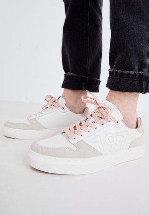 Baskets blanches et beiges avec des lacets et des accents roses. Comprend des matériaux en daim et en cuir lisse, avec un design de numéros sur le côté.