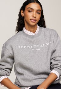 Felpa grigia con scollo rotondo, con un logo bianco che reca la scritta "TOMMY HILFIGER EST. 1985." Sovrapposta a una camicia bianca con righe rosse.