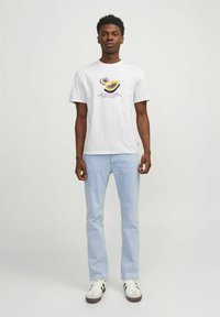 Witte katoenen t-shirt met een kleurrijk grafisch ontwerp met gele en paarse accenten, gedragen met lichtblauwe jeans en witte sneakers.