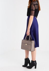 Borsa grigia con forma rettangolare, cuciture a contrasto e manici superiori. Modella che indossa un top in pizzo nero, una gonna a pieghe viola e stivali neri alla caviglia.