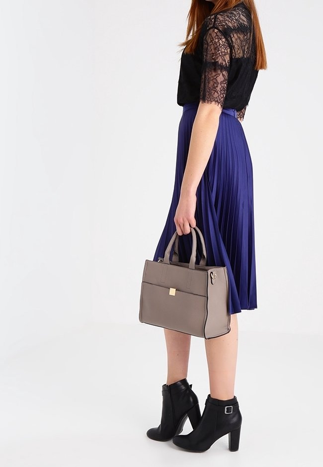 Borsa grigia con forma rettangolare, cuciture a contrasto e manici superiori. Modella che indossa un top in pizzo nero, una gonna a pieghe viola e stivali neri alla caviglia.