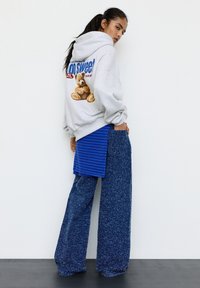 Felpa con cappuccio grigia con una grafica di un orsacchiotto sul retro, abbinata a pantaloncini a righe blu e pantaloni larghi in tessuto blu texturizzato.