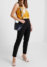 Blusa gialla Con motivo floreale, pantaloni neri slim-fit e sandali bianchi con tacco a blocco. La borsa è blu navy con texture coccodrillo.