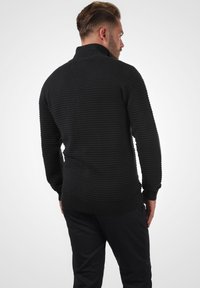 Schwarzer Strickpullover mit hohem Kragen, der über eine gerippte Textur und ein horizontales Streifenmuster verfügt, kombiniert mit dunklen Hosen.