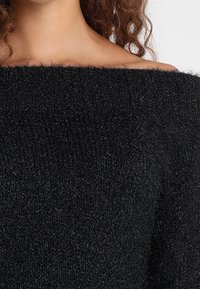 Czarny sweter odkrywający ramiona, wykonany z puchatej, teksturowanej tkaniny. Posiada szeroki dekolt i miękki, przytulny wygląd.