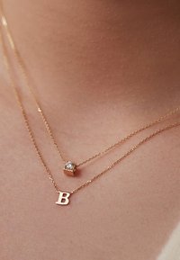 Gouden gelaagde halskettingen met een kubusvormige charm met een heldere steen en een hanger in de vorm van de letter "B". Delicaat kettingontwerp.