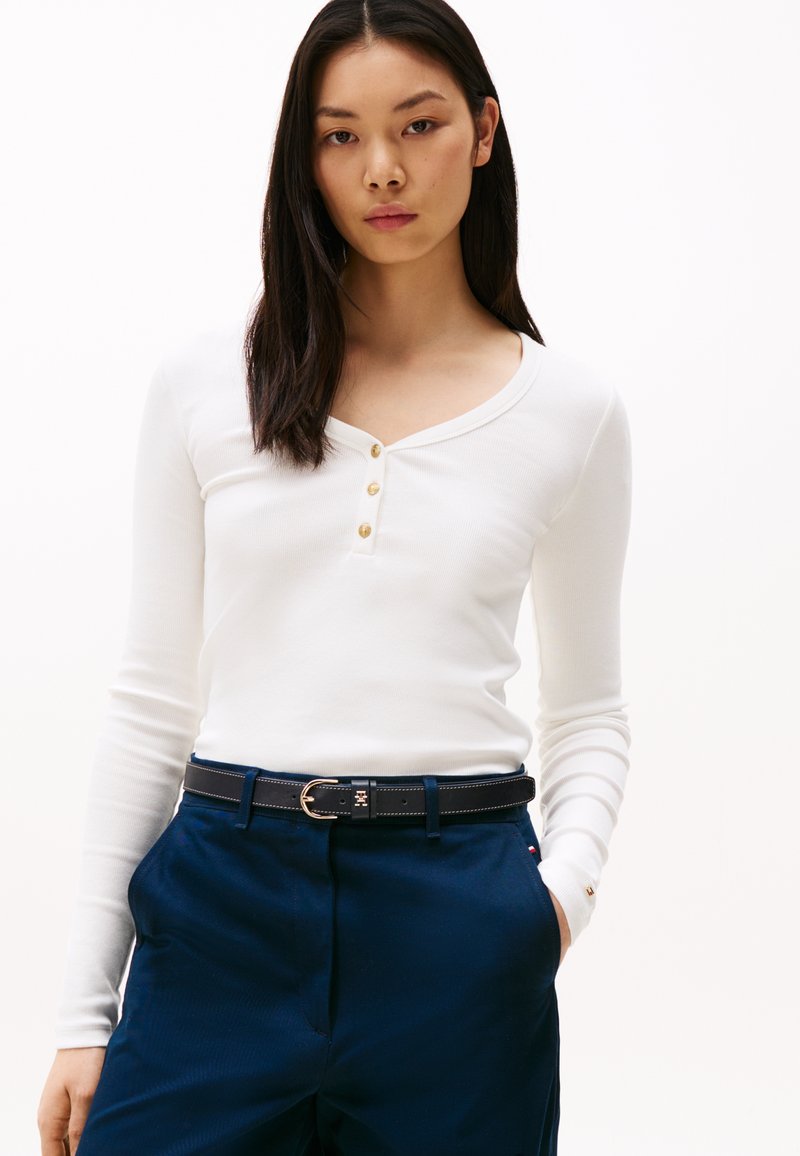 Long-sleeve white henley with three gold buttons, paired with navy trousers and a black belt featuring a gold buckle. Smooth texture.