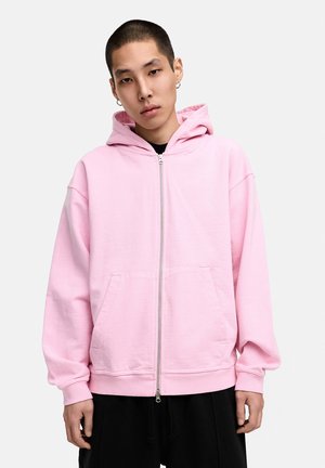 Helle rosa Kapuzenjacke mit durchgehendem Reißverschluss, einer vorderen Tasche und verstellbarem Kapuzenbereich, in lockerer Passform und mit gerippten Bündchen. Der Stoff wirkt weich und strukturiert.