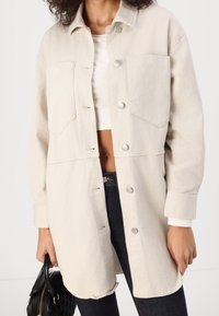 Person in light beige long jacket with silver buttons over white cropped top and dark jeans, holding a black quilted handbag.