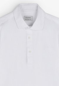 Weißes Poloshirt aus weichem Stoff, mit klassischem Kragen, drei Knöpfen und einem dezenten Logo auf der Brust. Schlichtes Design.
