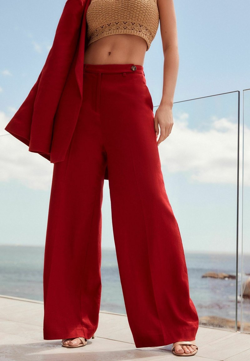 Femme portant un pantalon rouge à taille haute et jambes larges, un top court en crochet beige, tenant un blazer rouge assorti, debout près d'une rambarde en verre au bord de la mer.