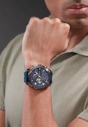 Moški z modno in srebrno zapestno uro z več merilniki, stisnjeno pest dvignjeno blizu obraza, oblečen v svetlo teksturirano polo majico.