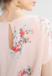 Blouse rose clair avec imprimé floral en rouge, bleu et vert. Présente un détail en découpe clé au dos et un tissu doux et léger. Manches courtes.