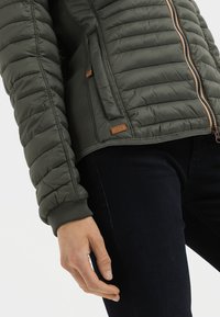 Gevoerde groene jas met een gladde textuur, horizontaal gestikt patroon, staande kraag, ritssluiting en bruin leren logo-detail.
