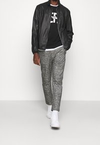 Svart läderjacka med hög krage, vit grafisk sweatshirt, grå texturerade byxor och vita sneakers i en gångställning.