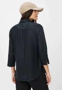 Dunkles, leichtes Hemd mit Dreiviertelärmeln, abgerundetem Saum und seitlichen Schlitzen. Kombiniert mit einer beigen Mütze, die ein lässiges Outfit zeigt.