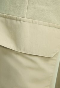 Beige tyg med en slät yta som har en horisontell söm och en fickklaff. Sömmen är synlig längs kanterna.