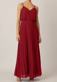 Femme portant une robe rouge plissée, sans manches, longueur au sol, avec des bretelles fines et des sandales à talons argentés sur un fond uni.