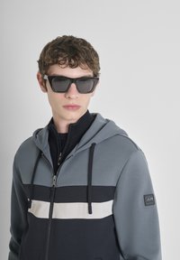 La felpa grigia con zip presenta un design a blocchi di colore con strisce di un blu navy scuro e bianco. Il modello indossa occhiali da sole quadrati scuri, sullo sfondo di una superficie semplice.