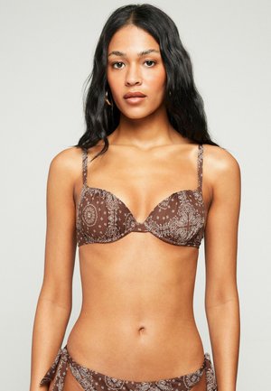 Femme aux longs cheveux noirs portant un haut de bikini marron à motifs et un bas assorti, debout devant un fond clair uni.