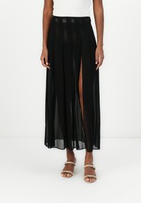 Gonna maxi nera a plissettature con pannelli trasparenti, caratterizzata da uno spacco laterale alto fino alla coscia e una vita con accenti a righe orizzontali. Texture liscia.