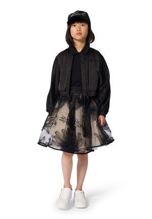 Zwarte satijnen bomberjack, zwart topje, beige tule rok met zwarte bloemenpatronen, witte sneakers en een zwarte pet met witte letters.