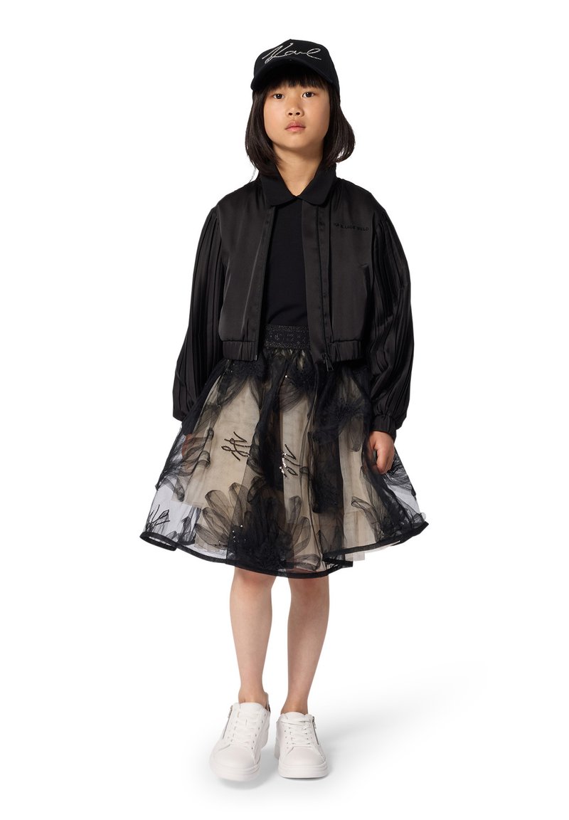 Veste bomber en satin noir, haut noir, jupe en tulle beige avec motifs floraux noirs, baskets blanches et casquette noire avec inscription blanche.