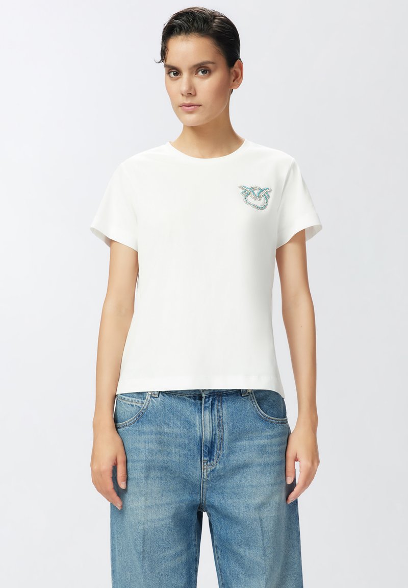 Camiseta blanca de manga corta con escote redondo. Presenta un diseño embellecido en turquesa en el área del pecho izquierdo. Material de algodón suave.