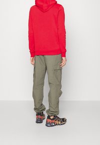 Sudadera roja con capucha, pantalones cargo oliva con bolsillos laterales y zapatillas negras con detalles en naranja. Enfoque en diseño deportivo y casual.
