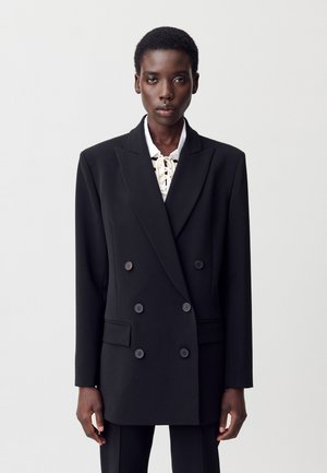 Blazer negro de corte doble con solapas afiladas, hombros estructurados y dos bolsillos delanteros. Presenta botones grandes y una textura suave.