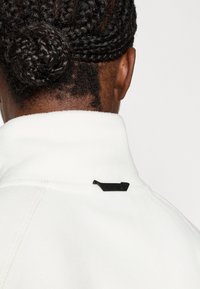 Λευκή ζακέτα από φλις με υψηλό γιακά, διαθέτει μαύρη ετικέτα στο πίσω μέρος. Η υφή φαίνεται απαλή, με εμφανείς ραφές.