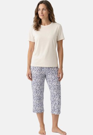 Kobieta stoi boso, ubrana w prostą beżową koszulkę z krótkim rękawem i przycięte spodnie w kwiatowy wzór na białym tle.