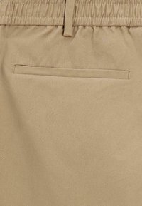 Béžová látka s elastickým pasem a plochým zadním kapsou. Textura je hladká s jemným leskem.