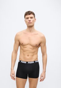 Νεαρός άνδρας με κοντά σγουρά μαλλιά που φοράει μαύρα εσώρουχα Puma boxer, στέκεται μπροστά σε απλό φωτεινό φόντο με ουδέτερη έκφραση.