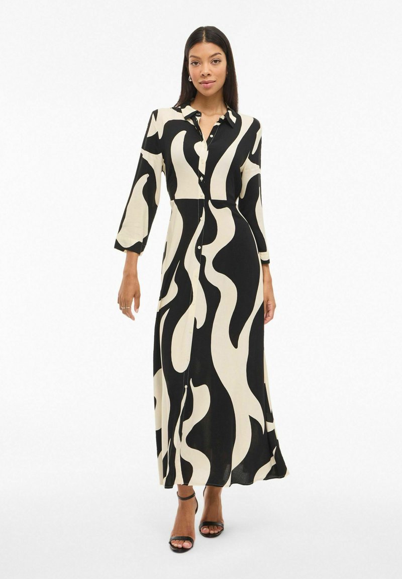 Dolga črna in smetana obleka z gumbnim zapenjanjem spredaj, ovratnikom, rokavi do treh četrtin in valovnim vzorcem, izdelana iz lahkega blaga.