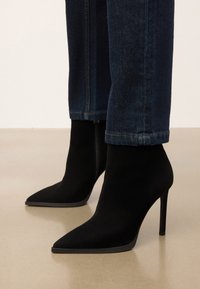 Mustad nahast terava ninaga stilettokontsadega põlvini saapad, mille juurde on kombineeritud tume sinine teksapüksid, asuvad beežil põrandal.