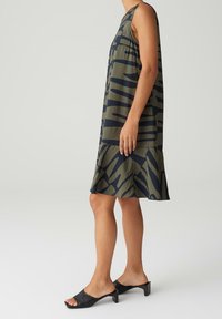 Robe sans manches longueur genoux avec un motif abstrait vert et noir portée par une personne debout avec des chaussures noires à talons bas et bouts ouverts.