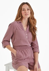 Woman with long wavy hair wearing a mauve textured button-up top and matching high-waisted shorts, seated with one hand in pocket.