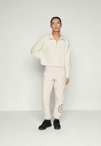 Pullover crema con colletto con zip e design cropped abbinato a pantaloni jogger beige chiaro con stampa di un logo circolare nero; indossato con sneakers nere.